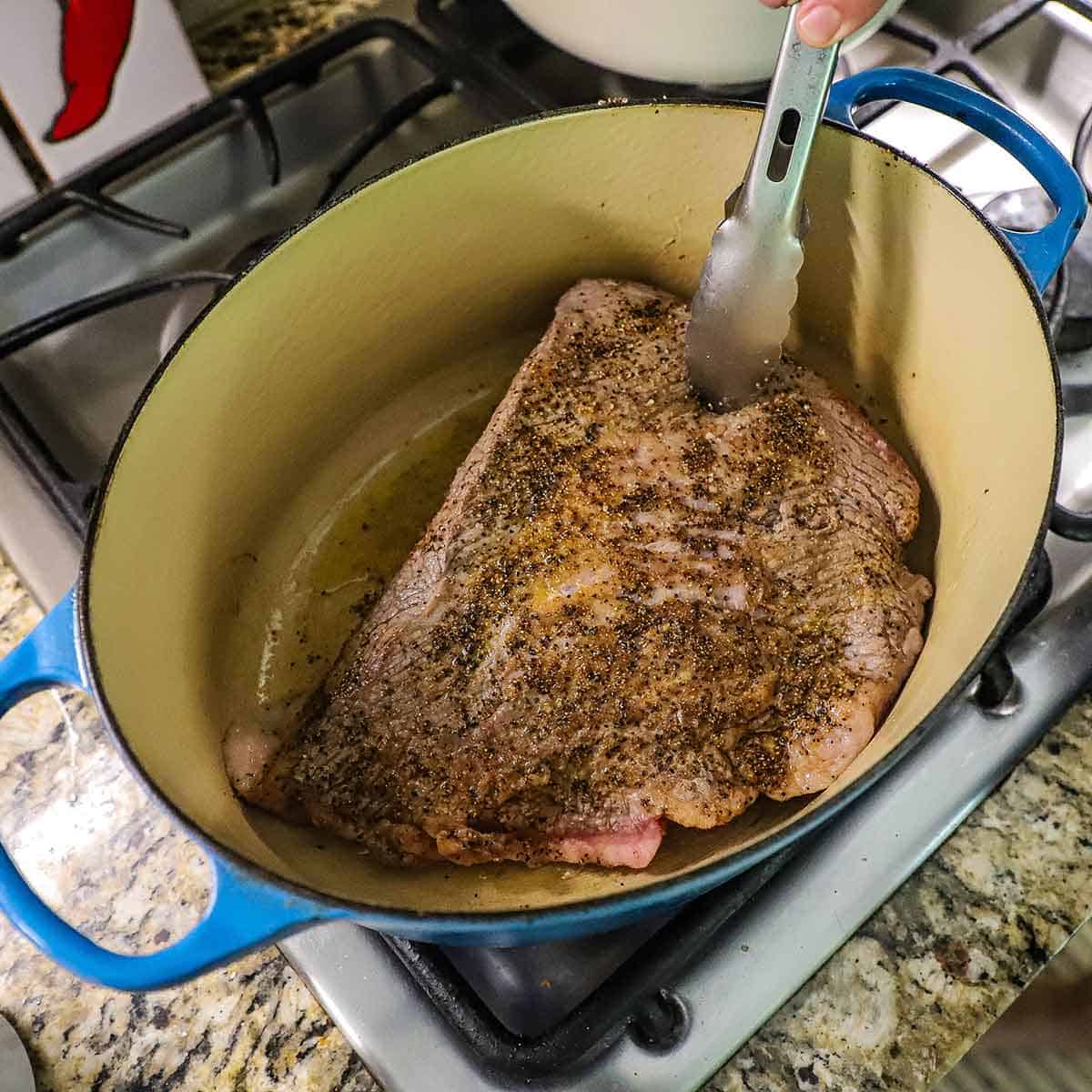 A person using a pair of tongs to hold up one side of a seasoned brisket flat that is being seared in an oval Dutch oven on a gas stove.