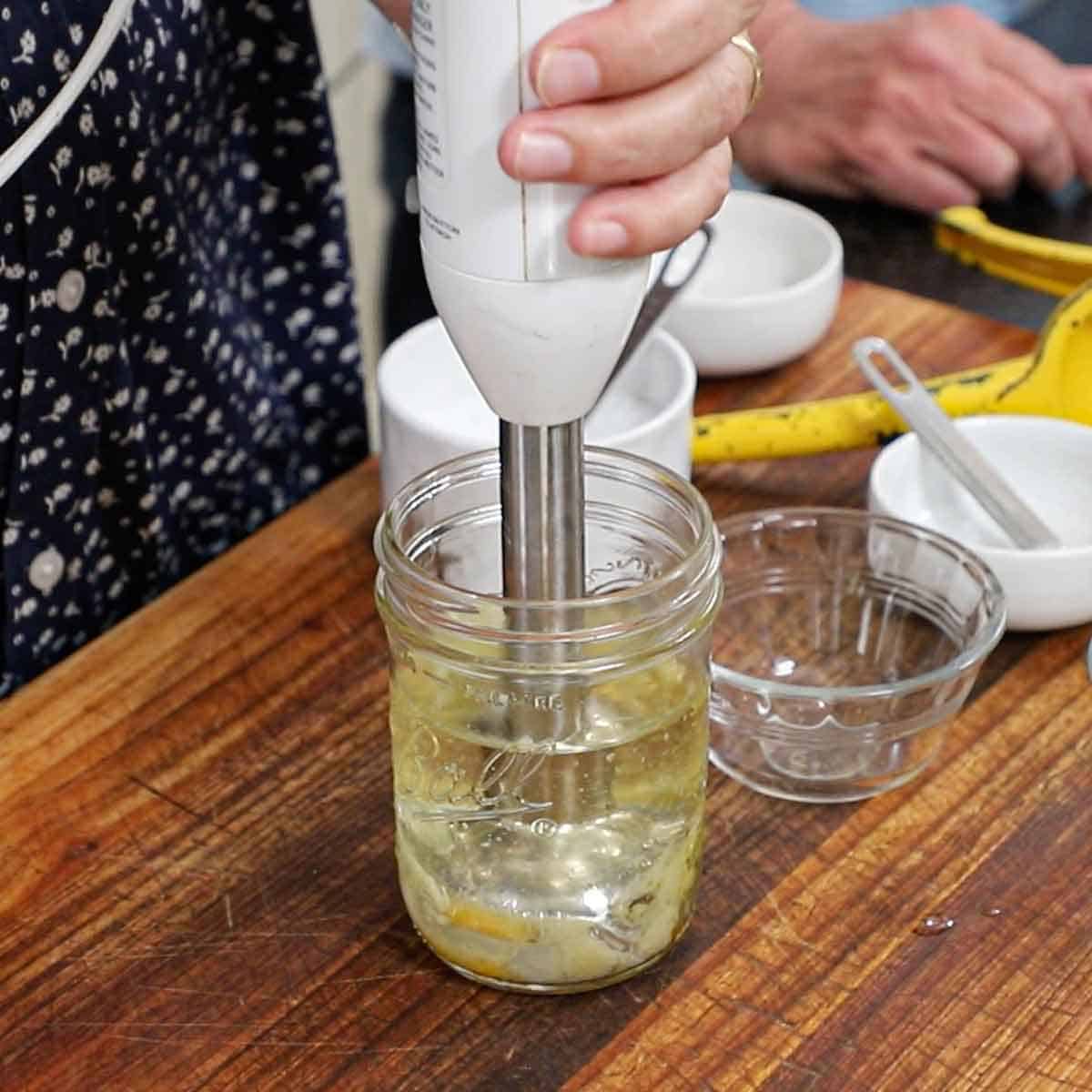 A person using an immersion blender to blend ingredients in a glass jar to make homemade mayonnaise.