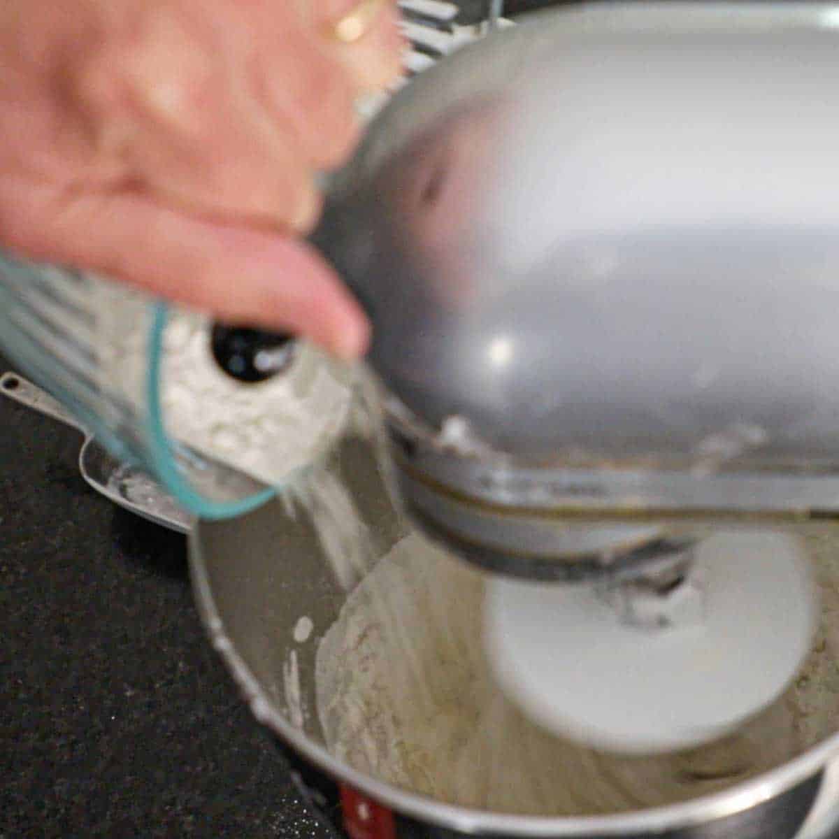 A person adding all-purpose flour into a stand-mixer that is beating dough for pepperoni rolls.