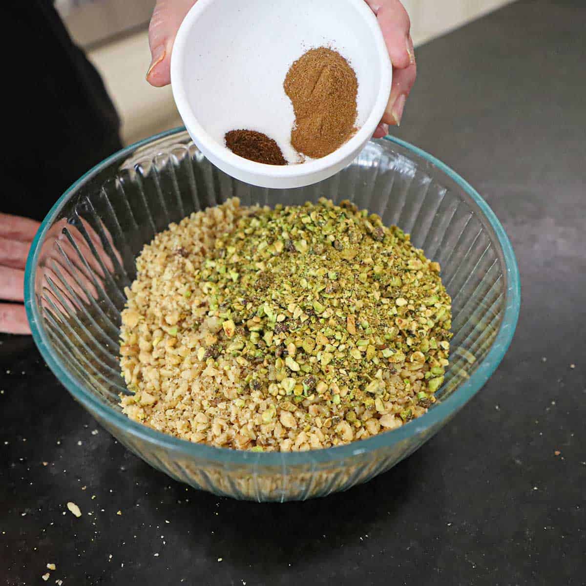A person transferring ground cinnamon, ground cloves, and salt from a small white bowl into a large glass bowl filled with pulverized walnuts and pistachios.