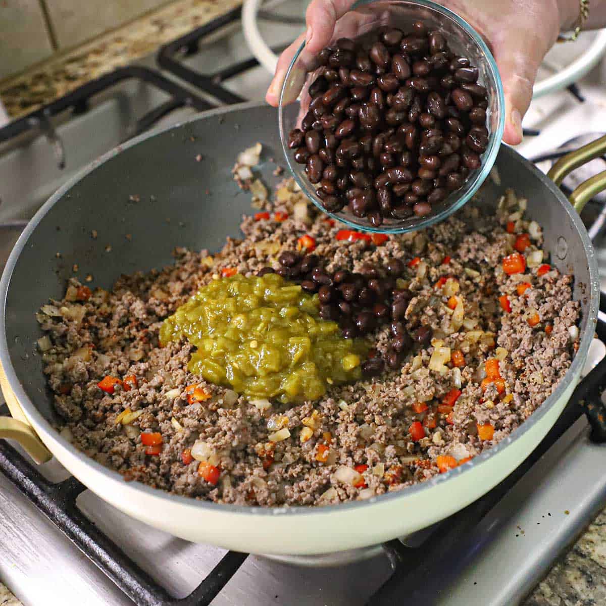 A person transferring black beans from a glass bowl into a skillet that is filled with cooked ground beef, saut&eacute;d onions and peppers, and diced green chiles.
