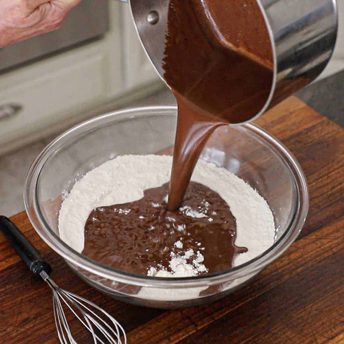 A person pouring a chocolate butter liquid from a saucepan into a large glass bowl filled with flour that has been mixed with sugar, baking soda, and salt.