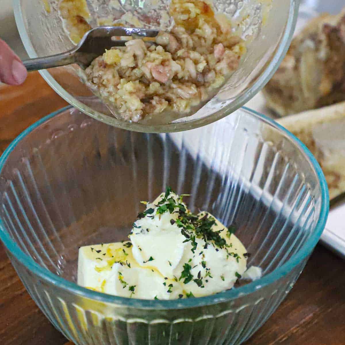 A person adding mashed roasted garlic and tender beef bone marrow from a small glass bowl into another bowl filled with softened butter, lemon zest, and fresh herbs.