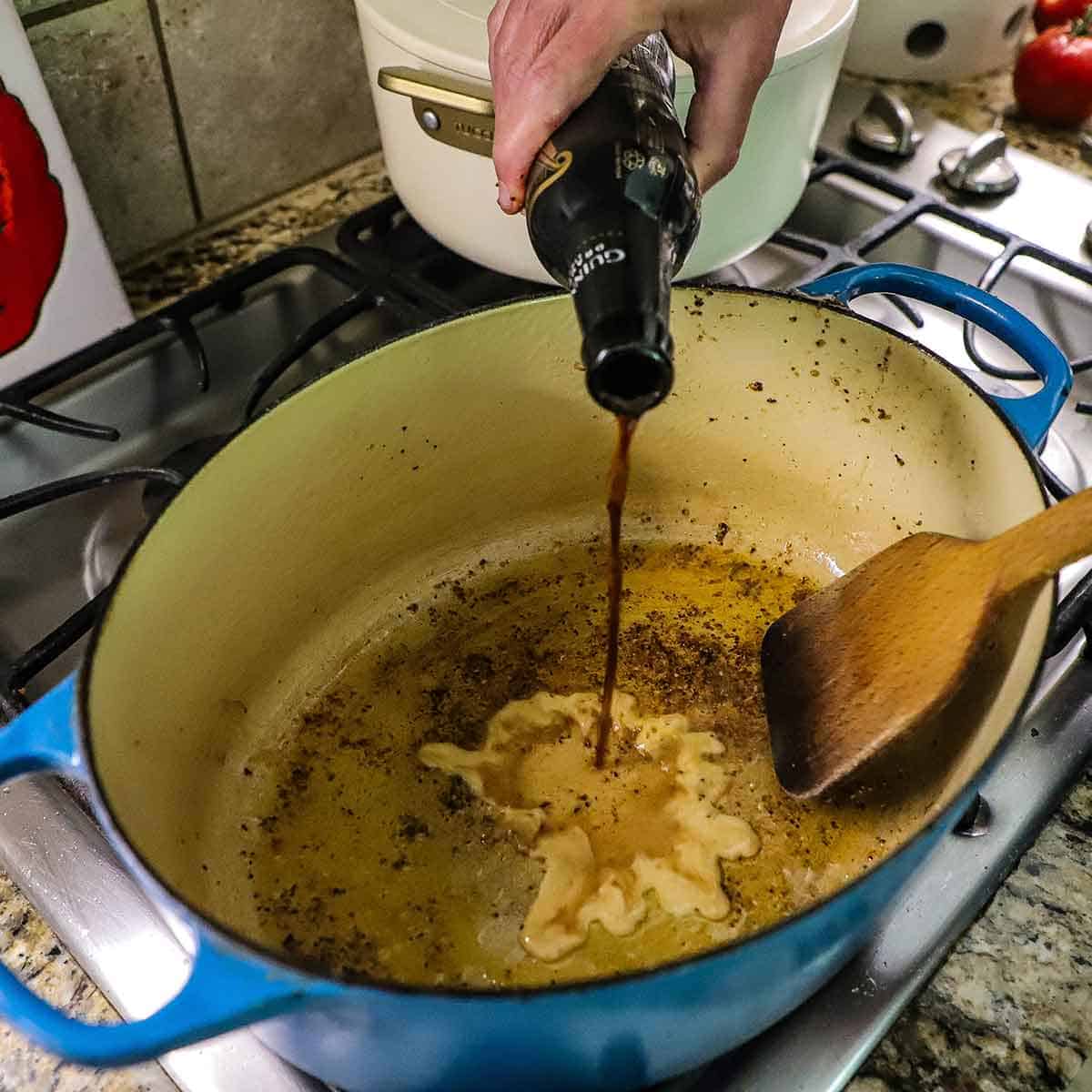 A person pouring a stout beef from a bottle into a Dutch oven that he is also using a wooden spatula to scrape up browned bits at the bottom of the pan.