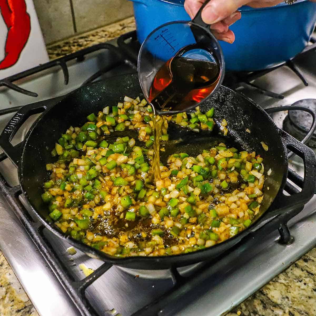 A person pouring bourbon from a clear measuring cup into a cast-iron skillet on a gas stove filled with simmering, sautéed onions, bell pepper, and garlic.