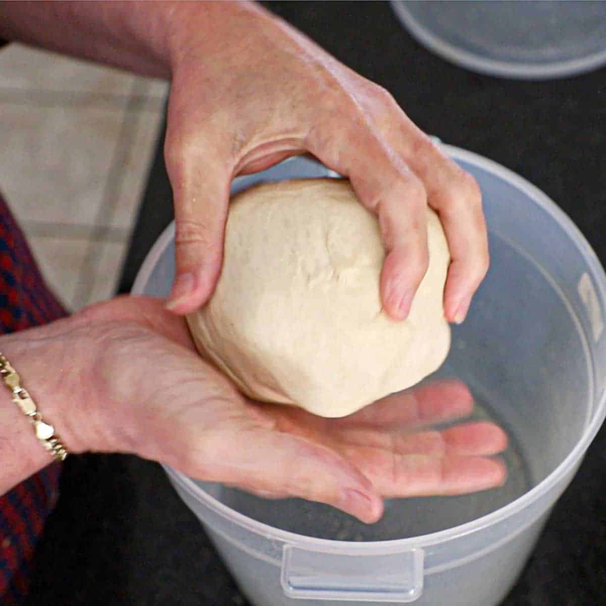 A person holding a smooth ball of pepperoni roll dough over a plastic proofing container.