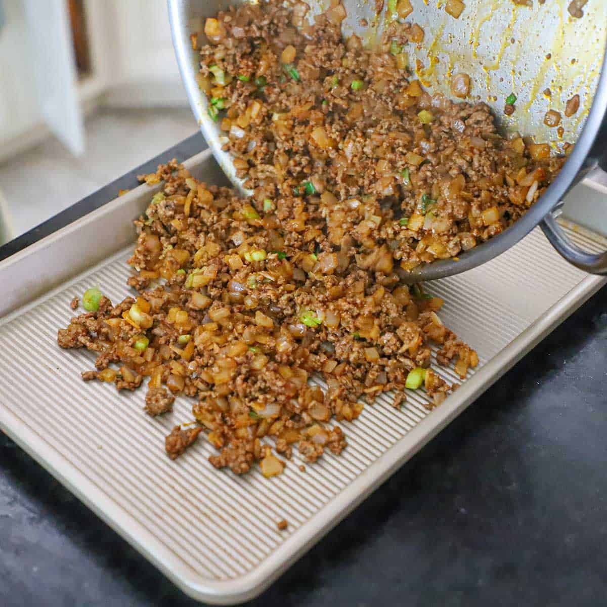 A person transferring a beef empanada filling from a large skillet onto a baking pan.