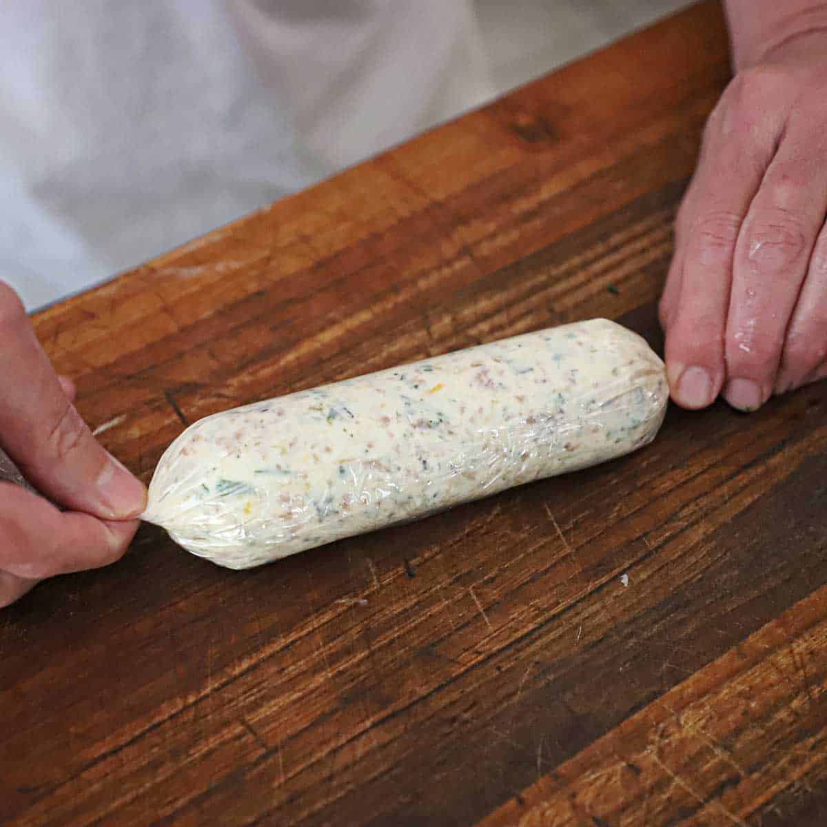 A person using his both hands to twist tight the ends of plastic wrap the have enclosed a soft log of roasted garlic and bone marrow compound butter.
