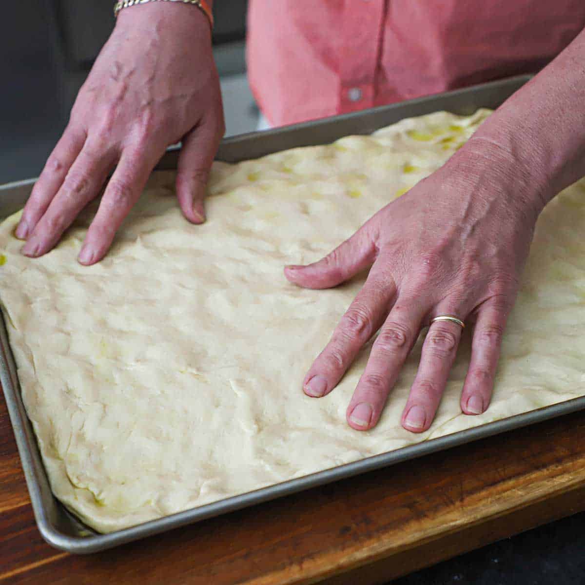 A person using his fingers and hands to stretch sheet pan pizza dough into a half sheet pan on a wooden cutting board.