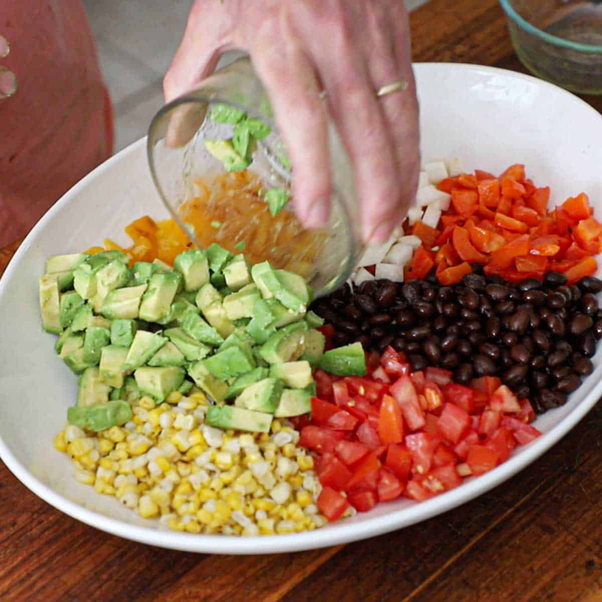 A person transferring cubed pieces of avocado into a large platter filled with piles of grilled corn kernels, tomatoes, roasted red bell peppers, diced jicama, and black beans.