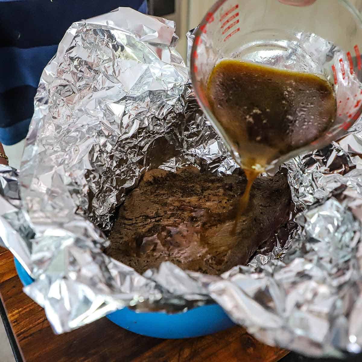 A person pouring a brown braising liquid from a glass measuring cup over a seared brisket flat that is nestled into two large sheets of foil inside a large Dutch oven.
