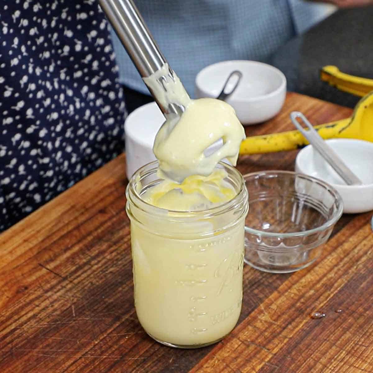 A person lifting an immersion blender out of a jar that is holding freshly made mayonnaise.