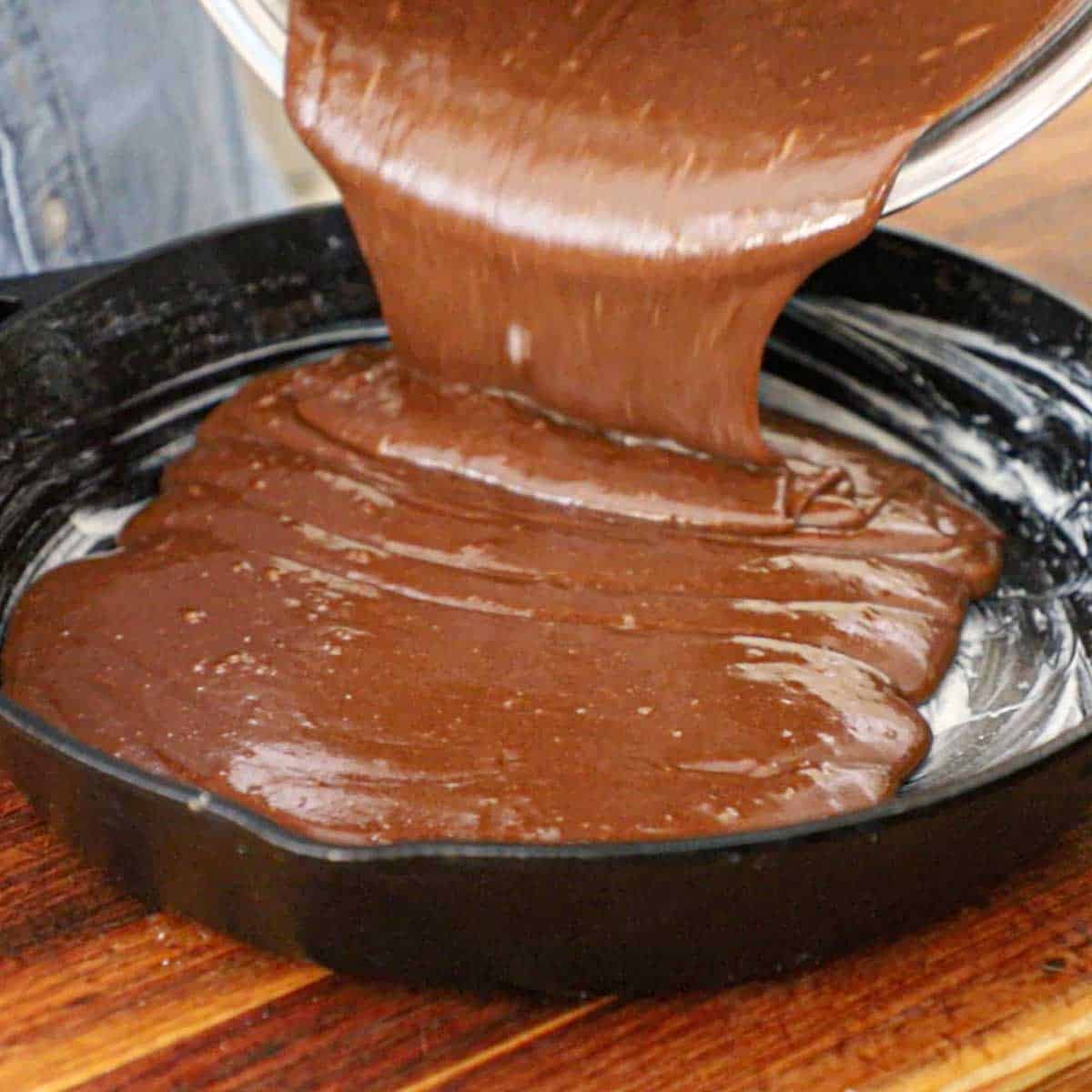 A person pouring a Texas Skillet Cake batter into a 12-inch cast-iron skillet that has been greased with softened butter.