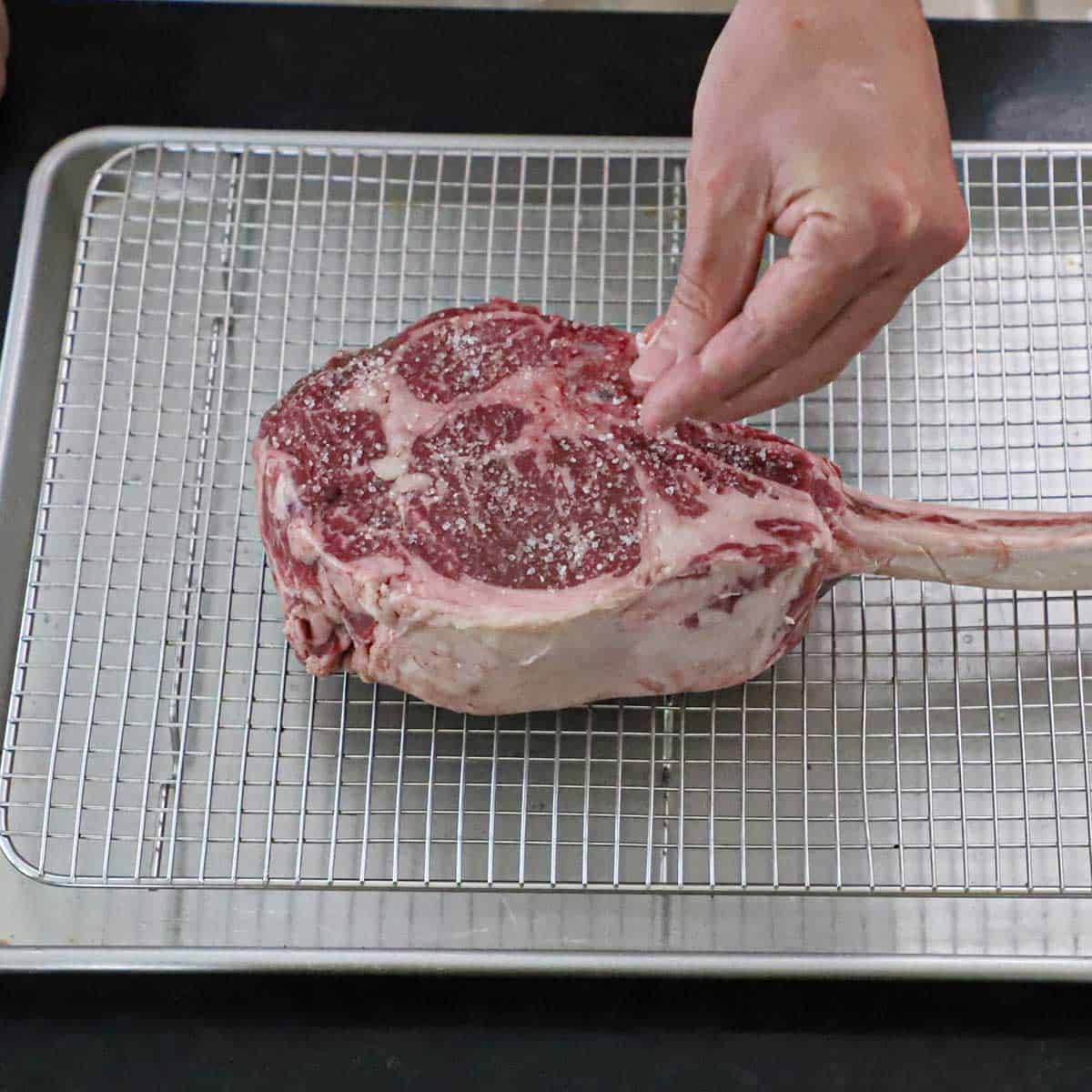 A person sprinkling salt over the surface of a tomahawk steak that is resting on a baking rack in a rimmed baking pan.