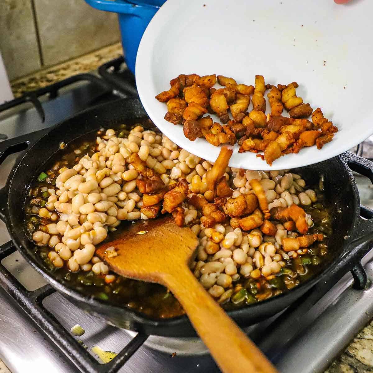 A person transferring crispy strips of pork belly from a white plate into a large cast-iron skillet on a gas stove filled with all the components of Bourbon-Glazed Baked Beans.