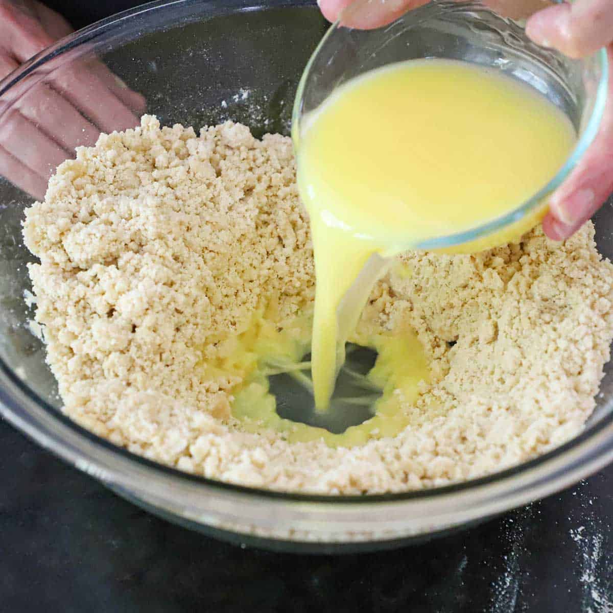 A person pouring an egg and water mixture into a bowl that is filled with a flour and pork lard mixture that has a well formed in the middle of the bowl.