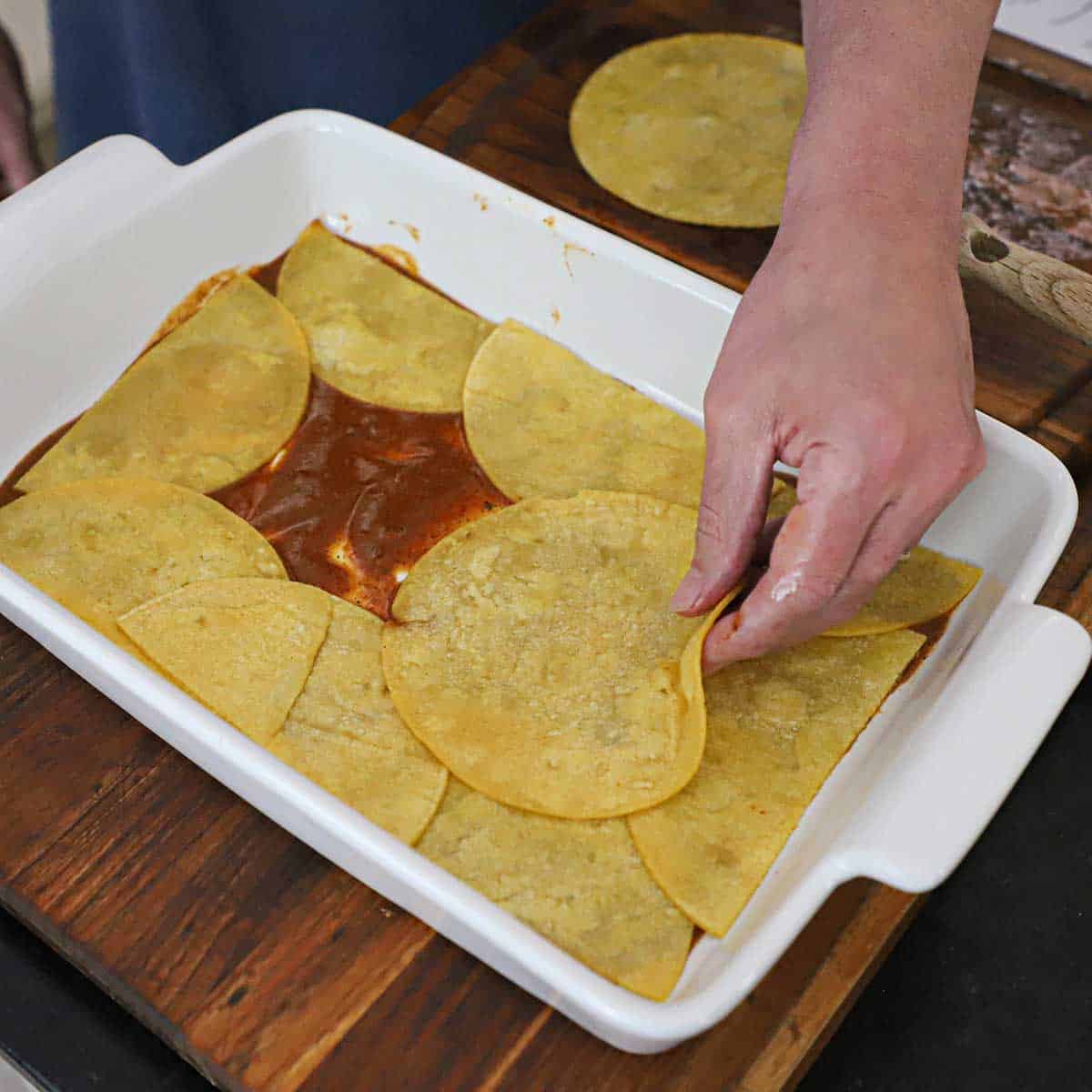 A person placing lightly fried corn tortillas into a 9 by 13-inch casserole dish with a thin layer of red enchilada sauce on the bottom of the dish underneath the tortillas.