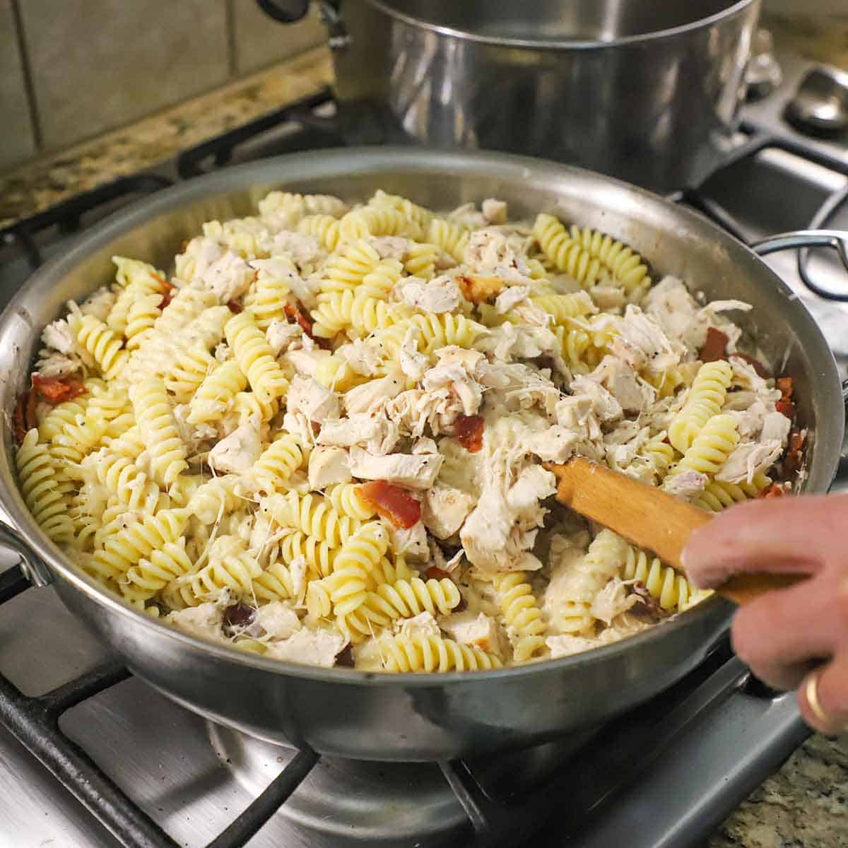 A person using a large wooden spoon to stir together cooked pasta, cooked chicken, bacon, and a creamy Mornay sauce in a large skillet on a gas stove.