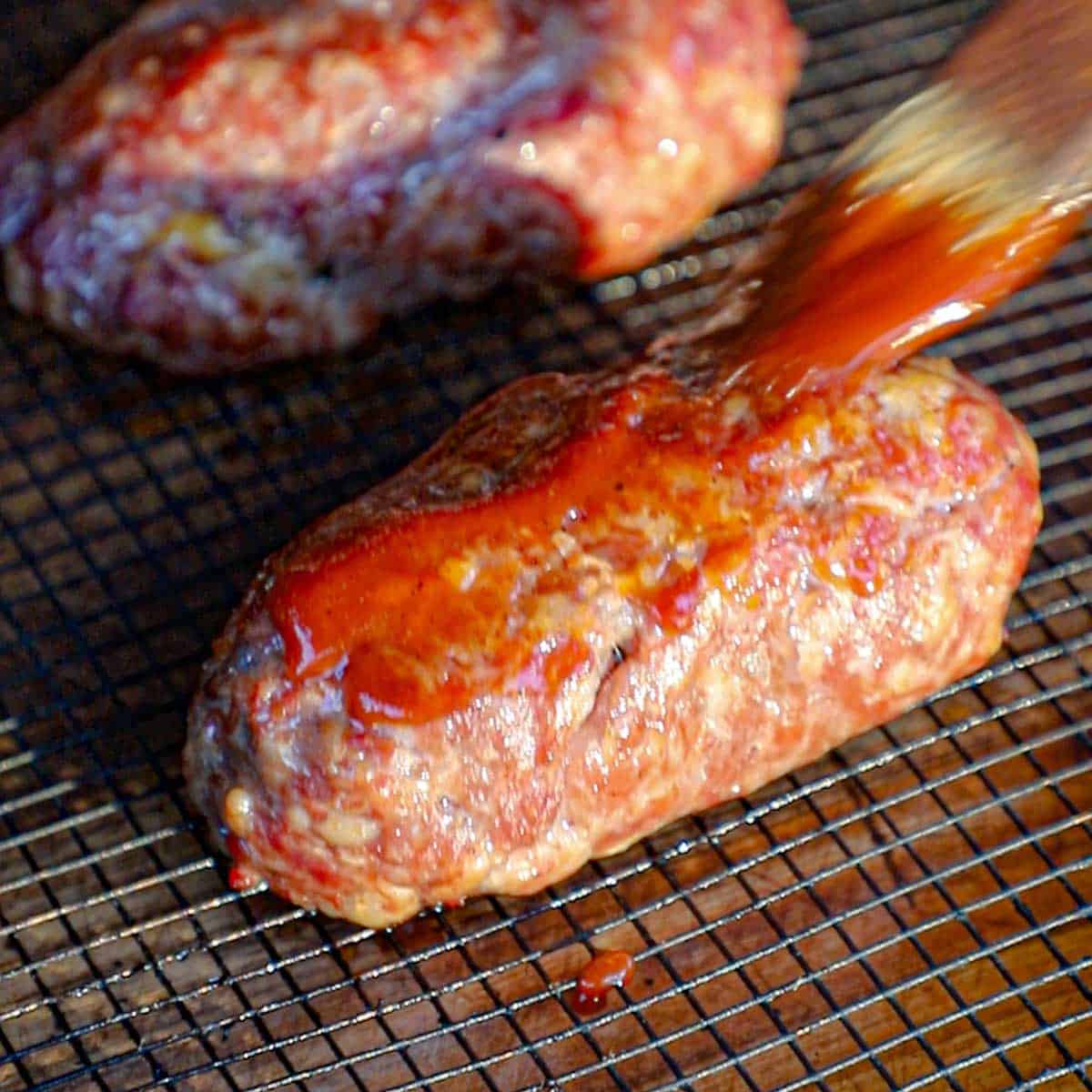 A person using a small brush to apply barbecue sauce onto a smoked armadillo egg that is resting on the grate of an electric smoker.