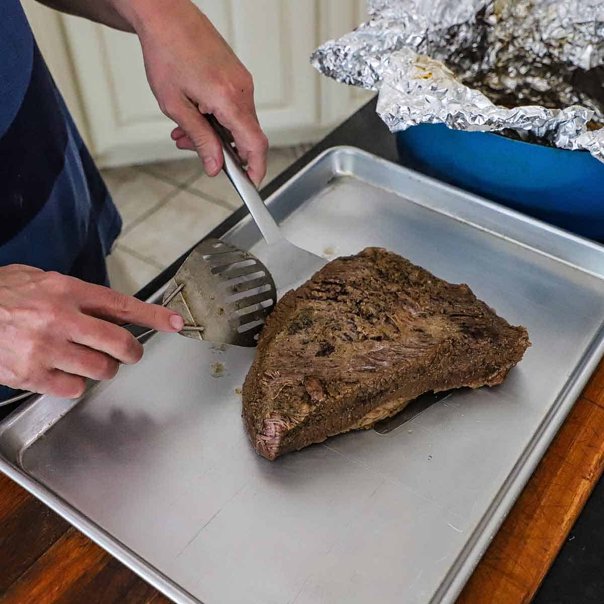 A person using a large spatula and spoon to place a fully cooked braised brisket flat on a large, rimmed sheet pan.