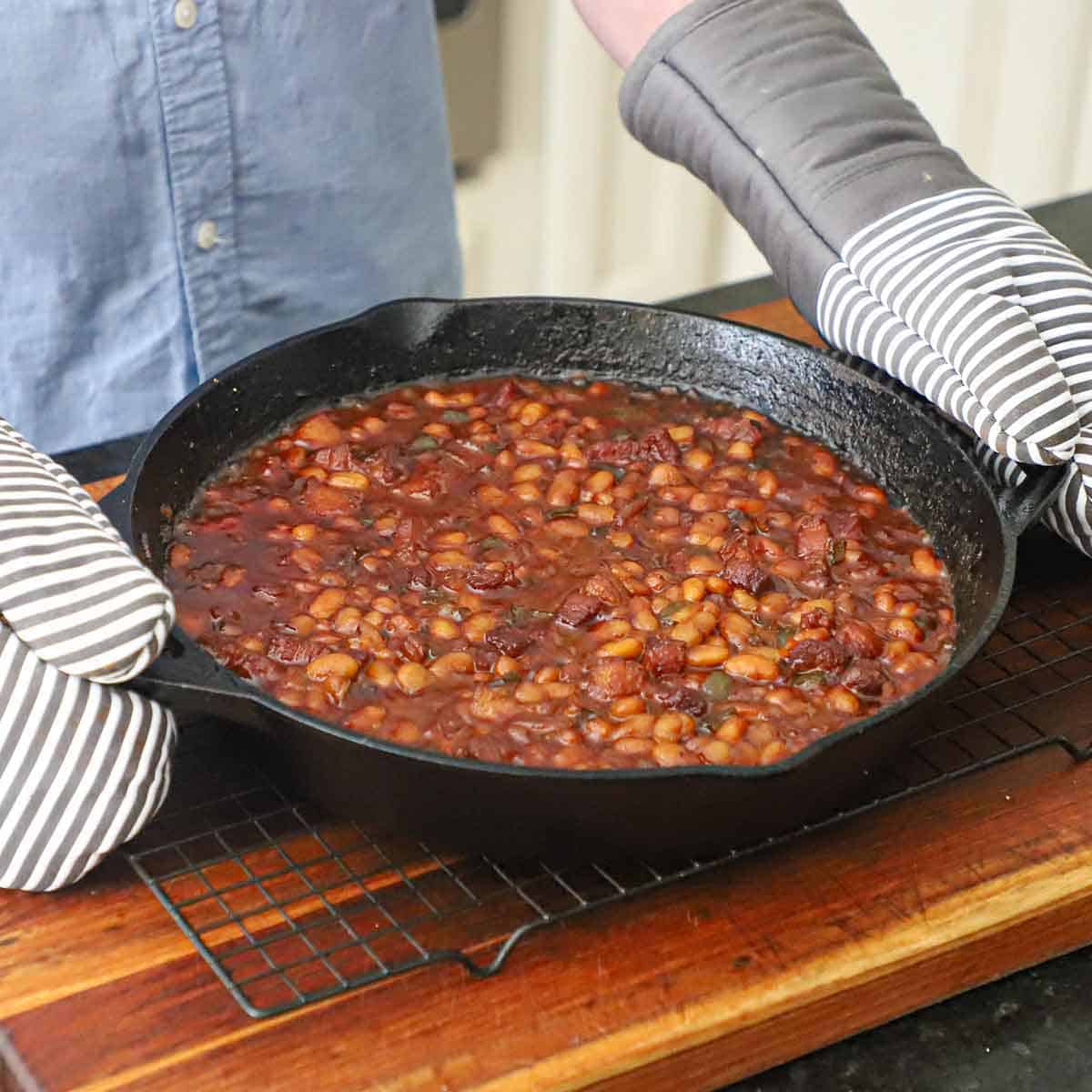 A person wearing oven mitts holding a large black cast-iron skillet that he has just removed from the oven and is filled with simmering Bourbon-Glazed Baked Beans with Pork Belly.