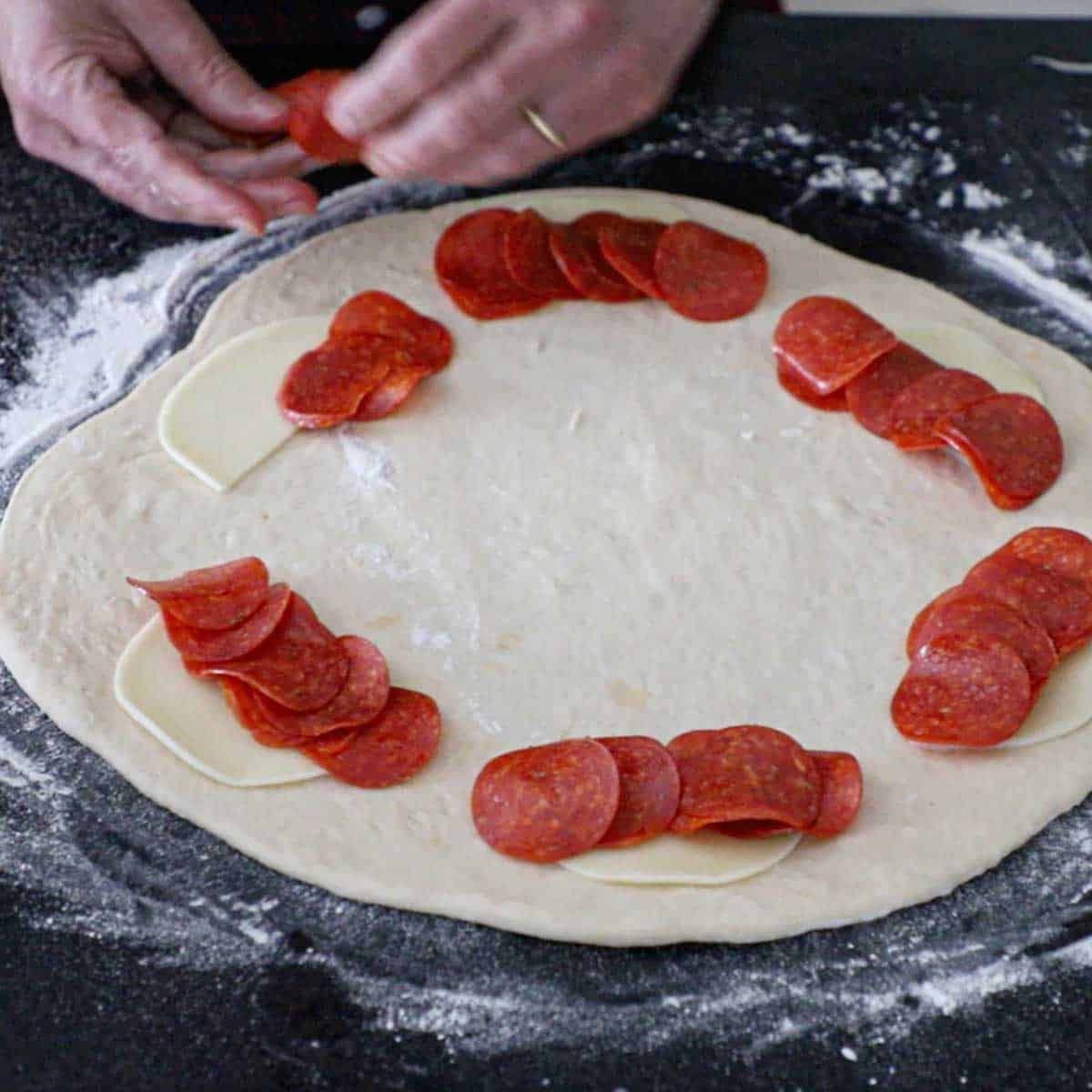 A person placing thinly sliced pepperoni over a strip of Provolone cheese along the out edges pepperoni roll dough that has been rolled into a 14-inch circle.