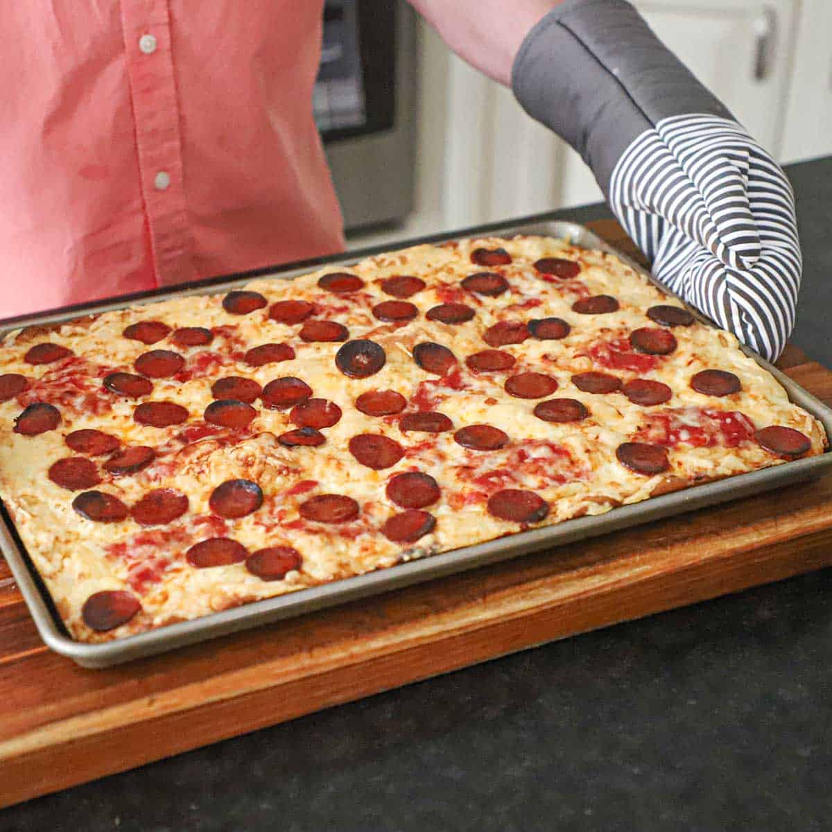 A person wearing oven mitts holding a homemade sheet pan pepperoni pizza that has just been removed from the hot oven.
