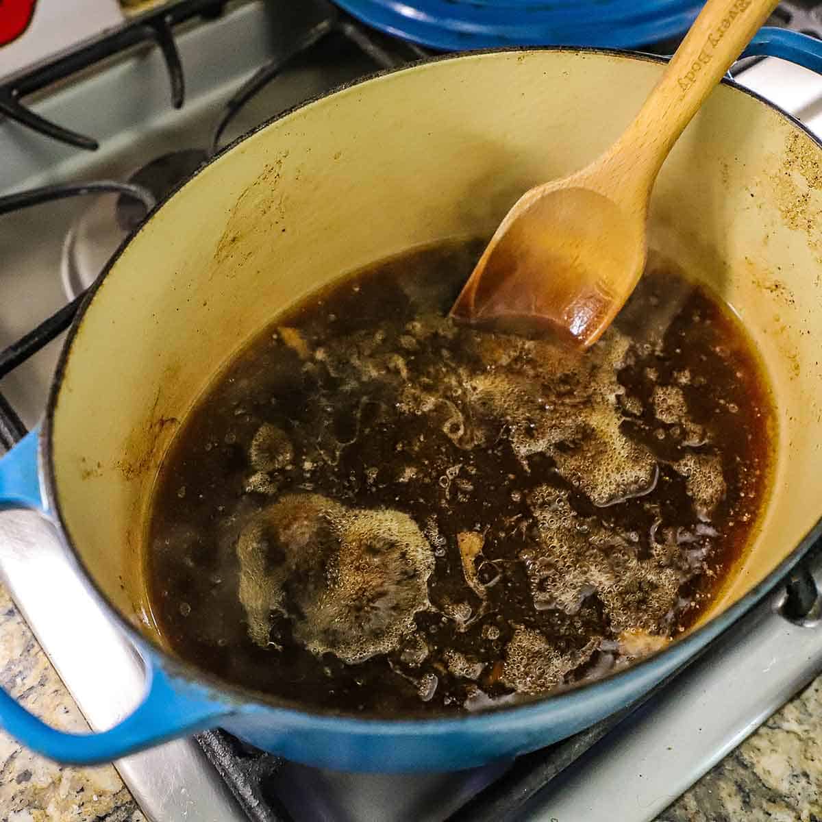 A person using a wooden spoon to stir simmering au jus sauce in a large Dutch oven on a gas stove.