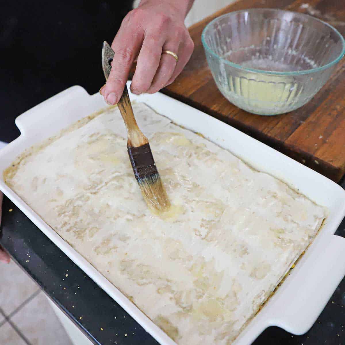 A person using a pastry brush to brush melted butter over the top of the top layer of baking dish filled with uncooked baklava.