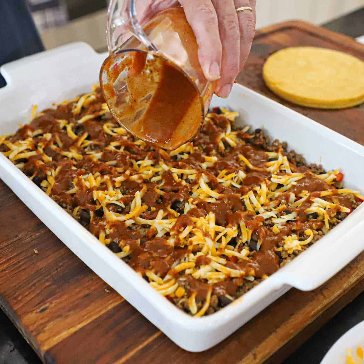A person pouring red enchilada sauce over the a layer of shredded cheese that is resting on top of a beef enchilada mixture in a 9 by 13-inch baking dish.