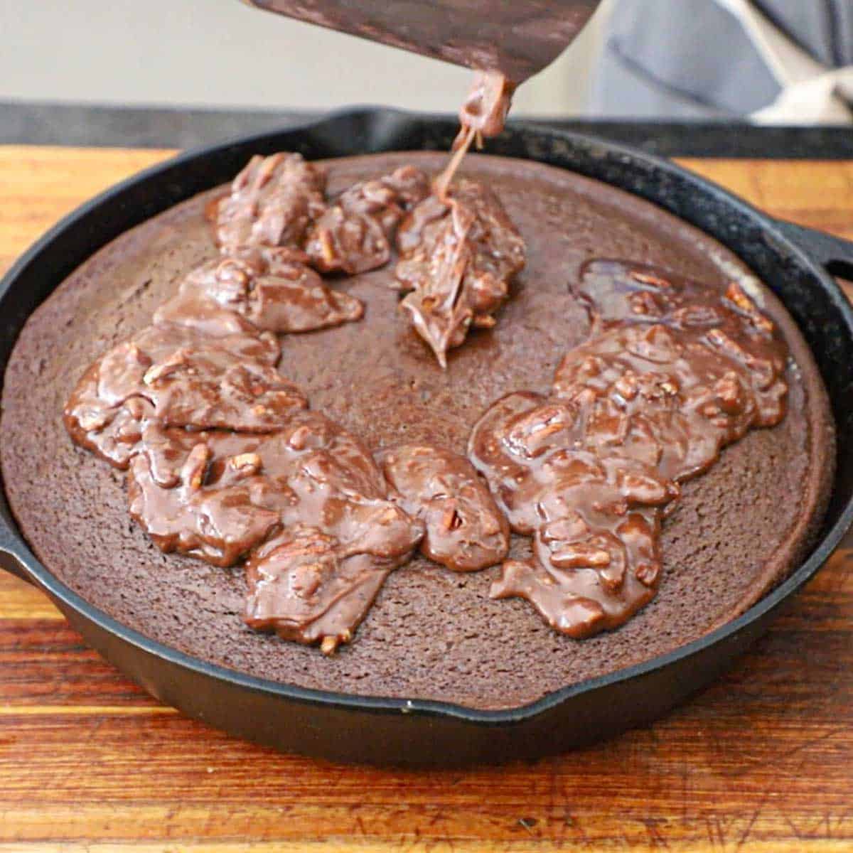 A person pouring a warm chocolate pecan frosting over the surface of a hot Texas Skillet Cake in a large cast-iron skillet.