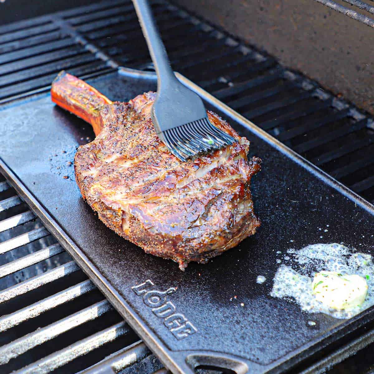 A person using a brush to apply melted bone marrow butter over a tomahawk steak that is being reverse seared on a cast-iron griddle on a gas stove.