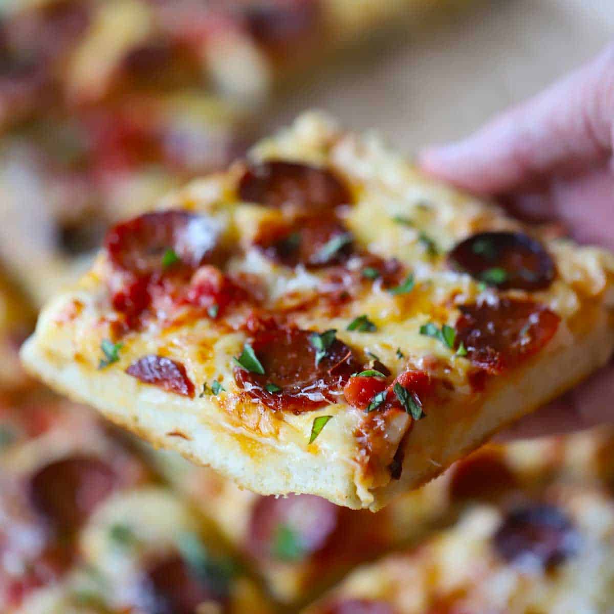 A person holding a square slice of a homemade sheet pan pepperoni pizza over the whole pizza below.