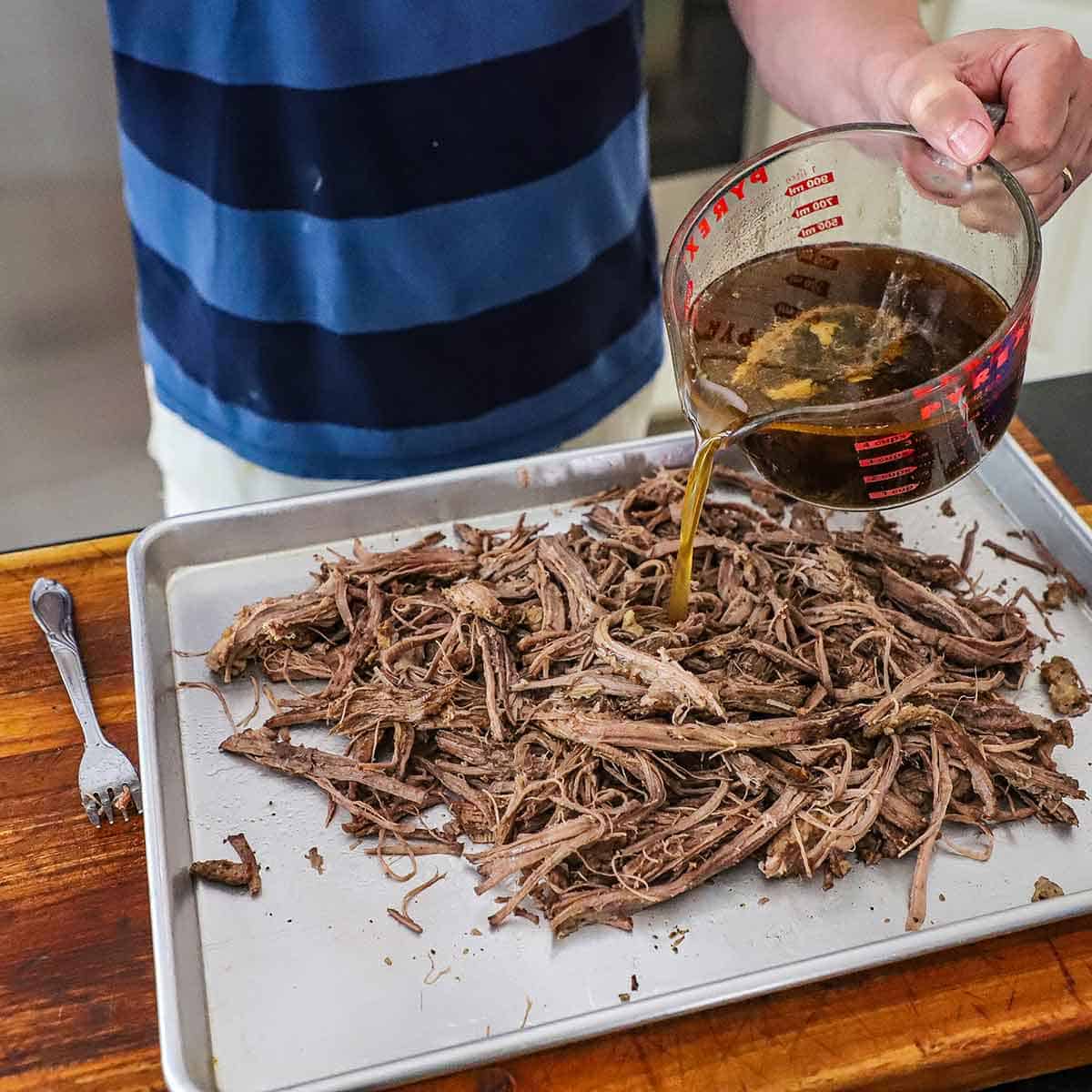 A person pouring an au jus sauce from a large glass measuring cup onto a large pile of shredded braised brisket on a rimmed sheet pan.