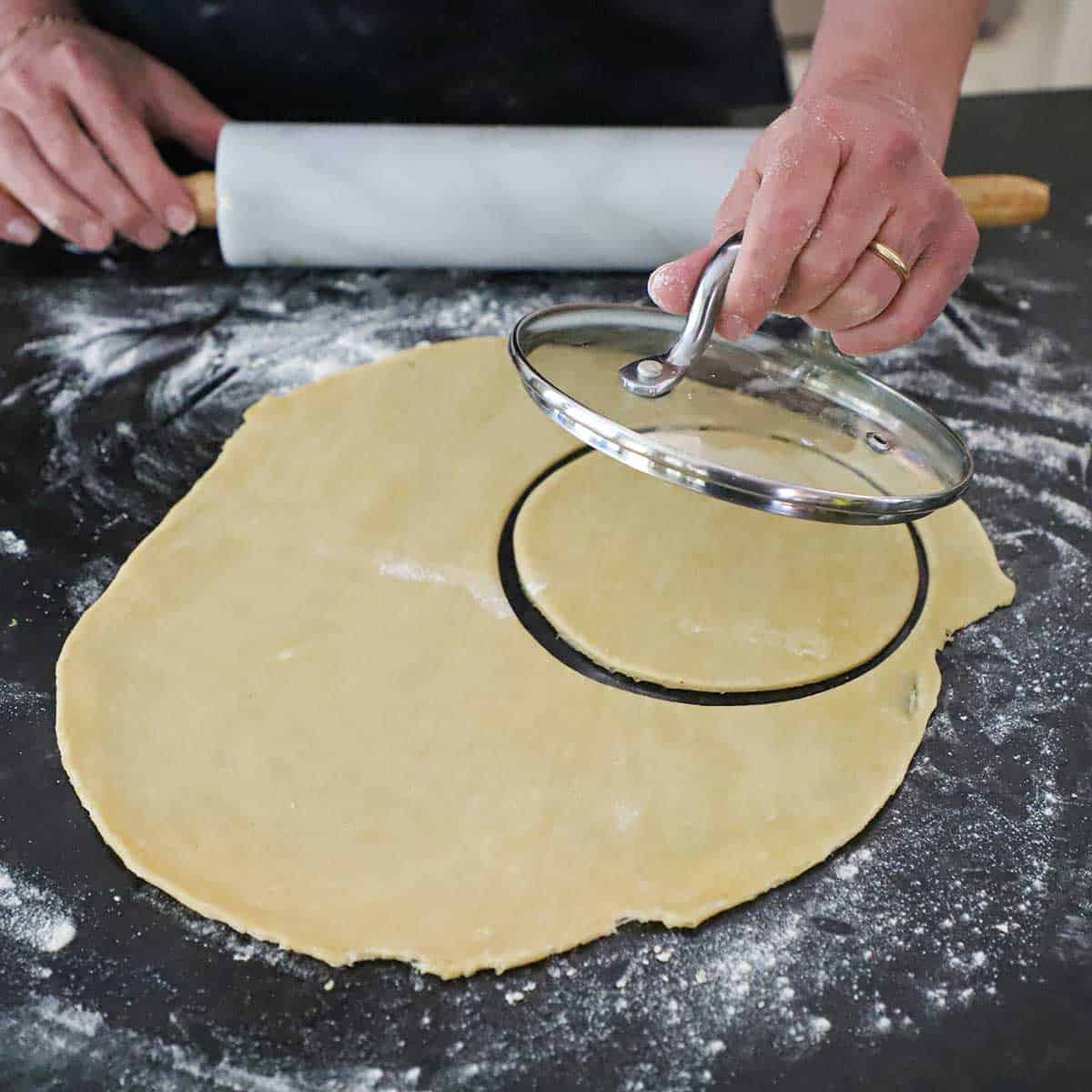 A person holding a glass top of a pan over a stretched out circular piece of empanada dough that he has used to cut a circular piece of dough.