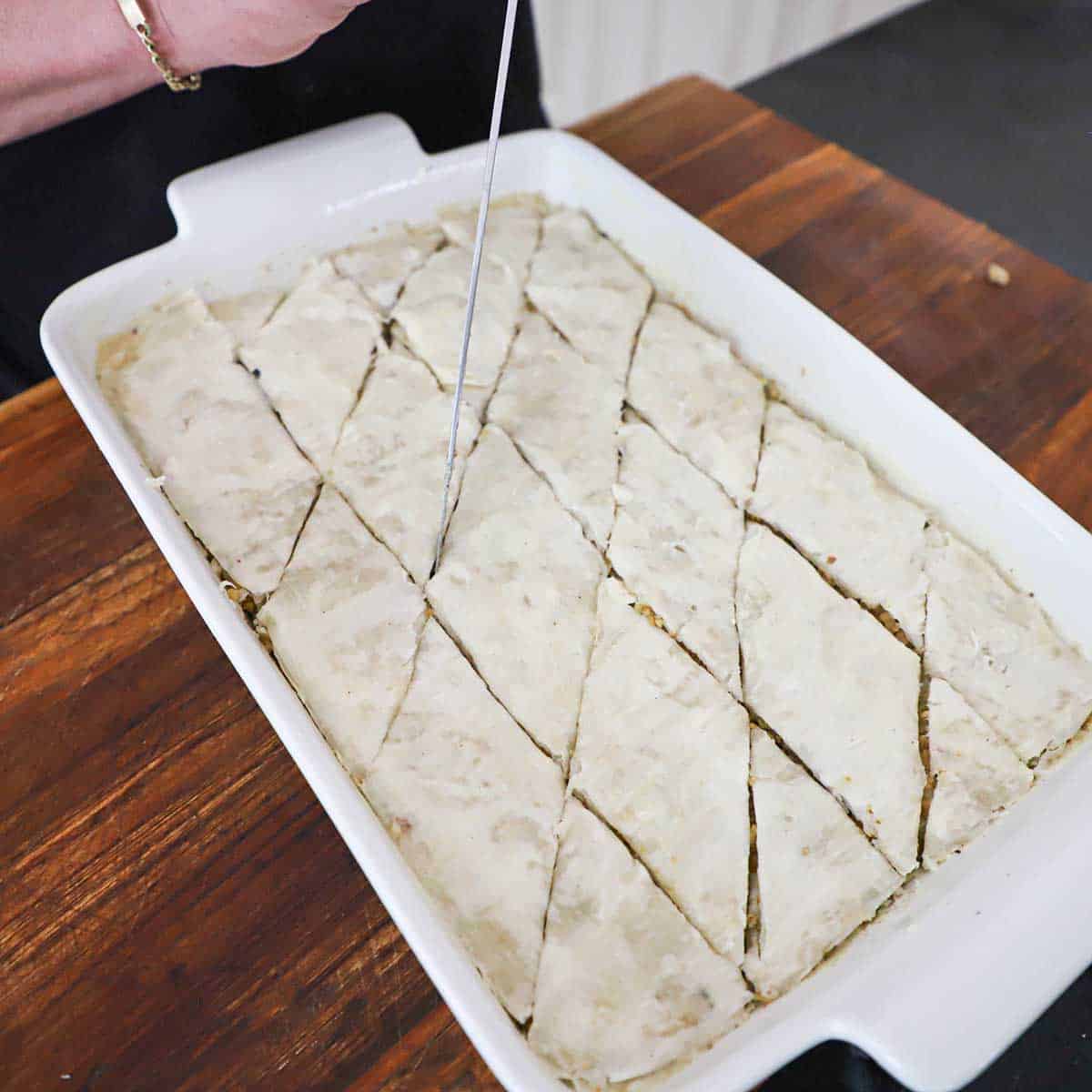 A person using a large chef's knife to slice unbaked baklava into diamond shapes in a 9x13" baking dish.