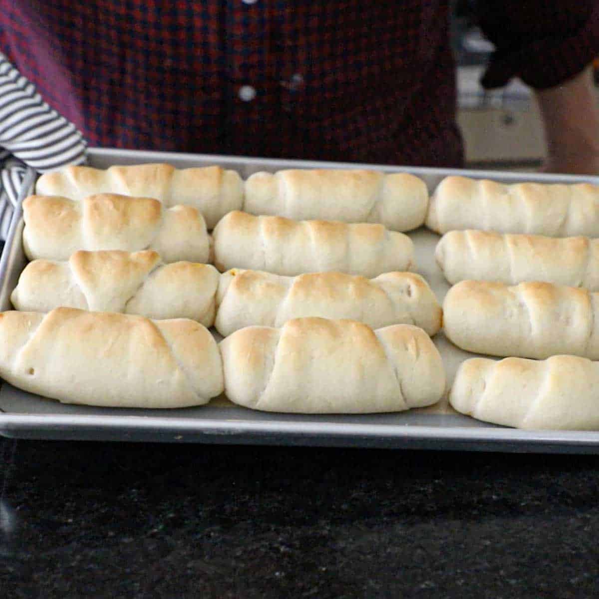 A person holding a large baking pan that is filled with 12 freshly baked Melt-in-Your-Mouth Pepperoni Rolls.