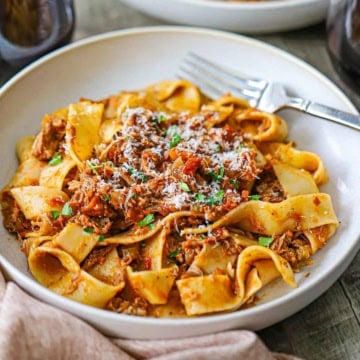 A close-up view of a shallow pasta bowl filled with a serving of Slow-Braised Pork Ragu with Pappardelle topped with finely shaved Parmesan cheese.