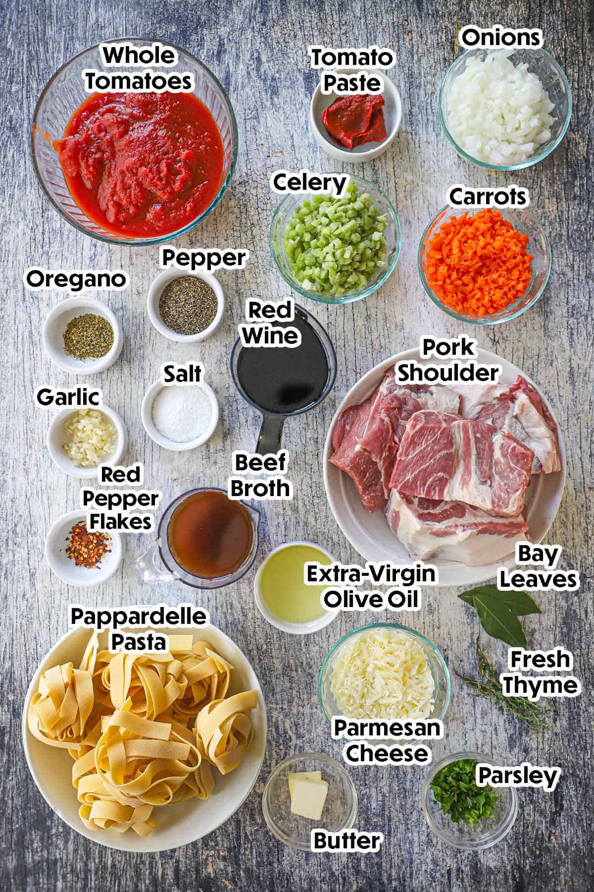 An arrangement of ingredients for Slow-Braised Pork Ragu with Pappardelle on a grey wooden background including pork shoulder, carrots, onions, celery, tomatoes, tomato paste, wine, garlic, oil, Parmesan cheese, butter, pasta, herbs, and seasonings.