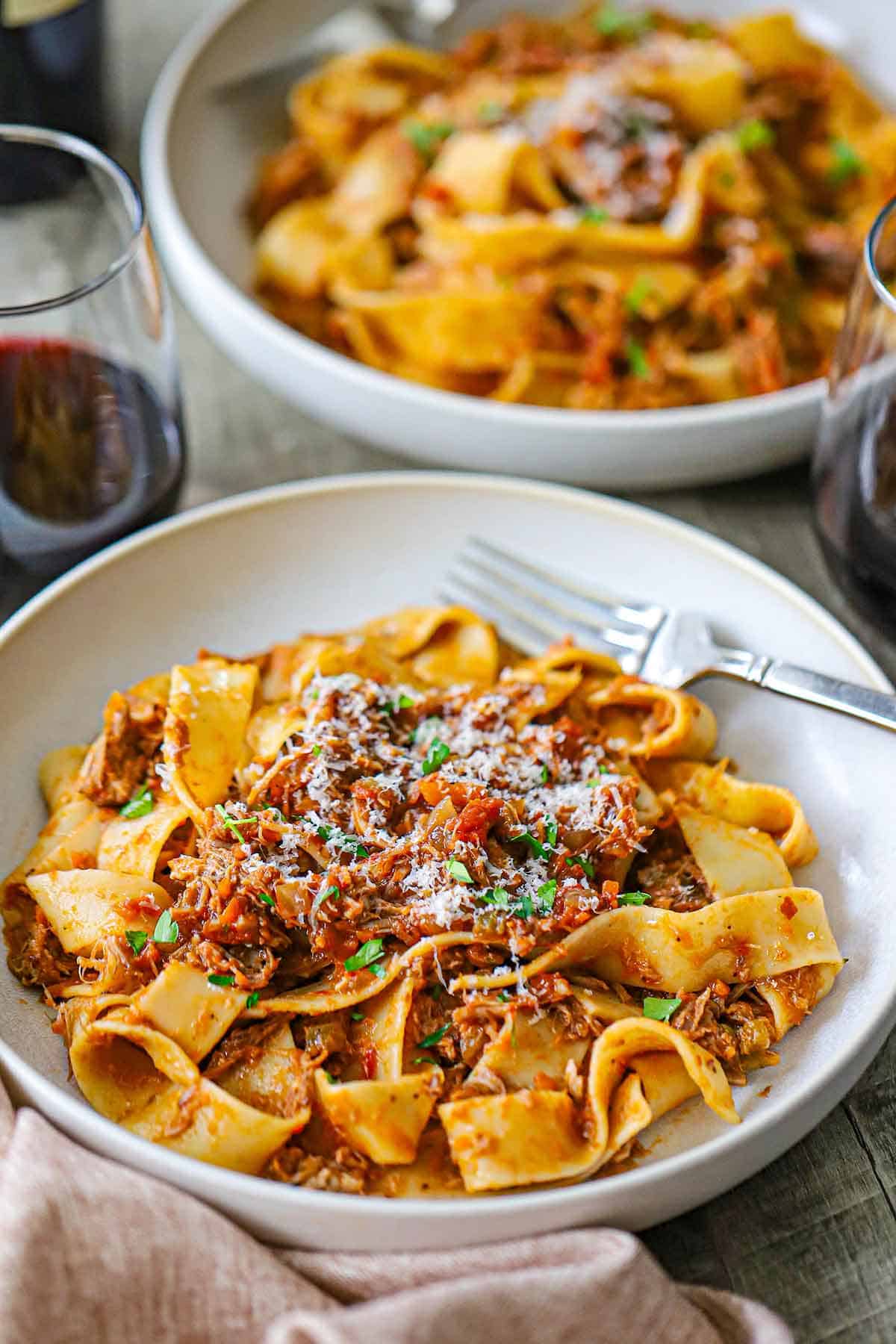 Two shallow pasta bowls filled with servings of Slow-Braised Pork Ragu with Pappardelle topped with finely shaved Parmesan cheese and two glasses of red wine near by.
