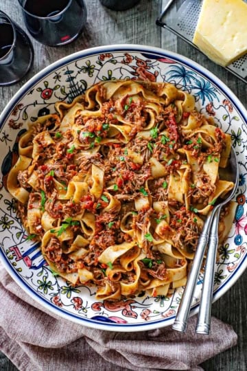 An overhead view of a large pasta bowl filled with Slow-Braised Pork Ragu with Pappardelle pasta with two silver serving spoons nestled into the side of the dish.