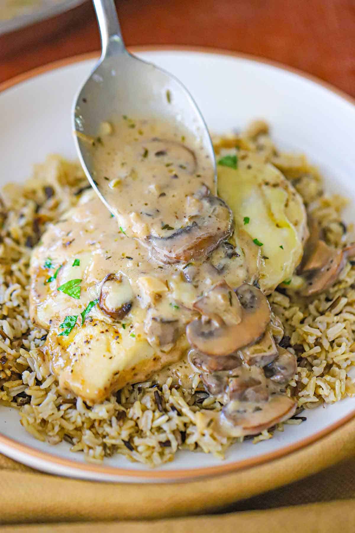 A person using a large spoon to drizzle a creamy mushroom sauce over the top of a seared chicken cutlet that is coated in melted cheese and is resting on a bed of wild rice in a shallow bowl.