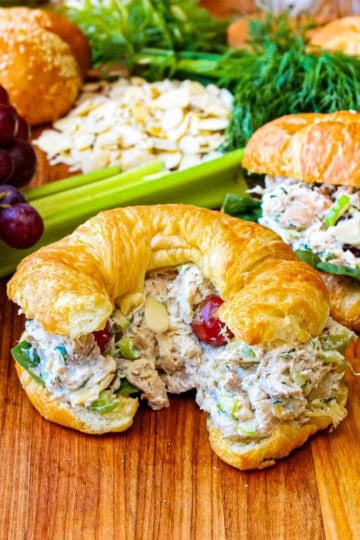 A croissant the is stuffed with Best-Ever Chicken Salad sitting on a cutting board next to another croissant also stuffed with the salad also resting near grapes, celery, and herbs.