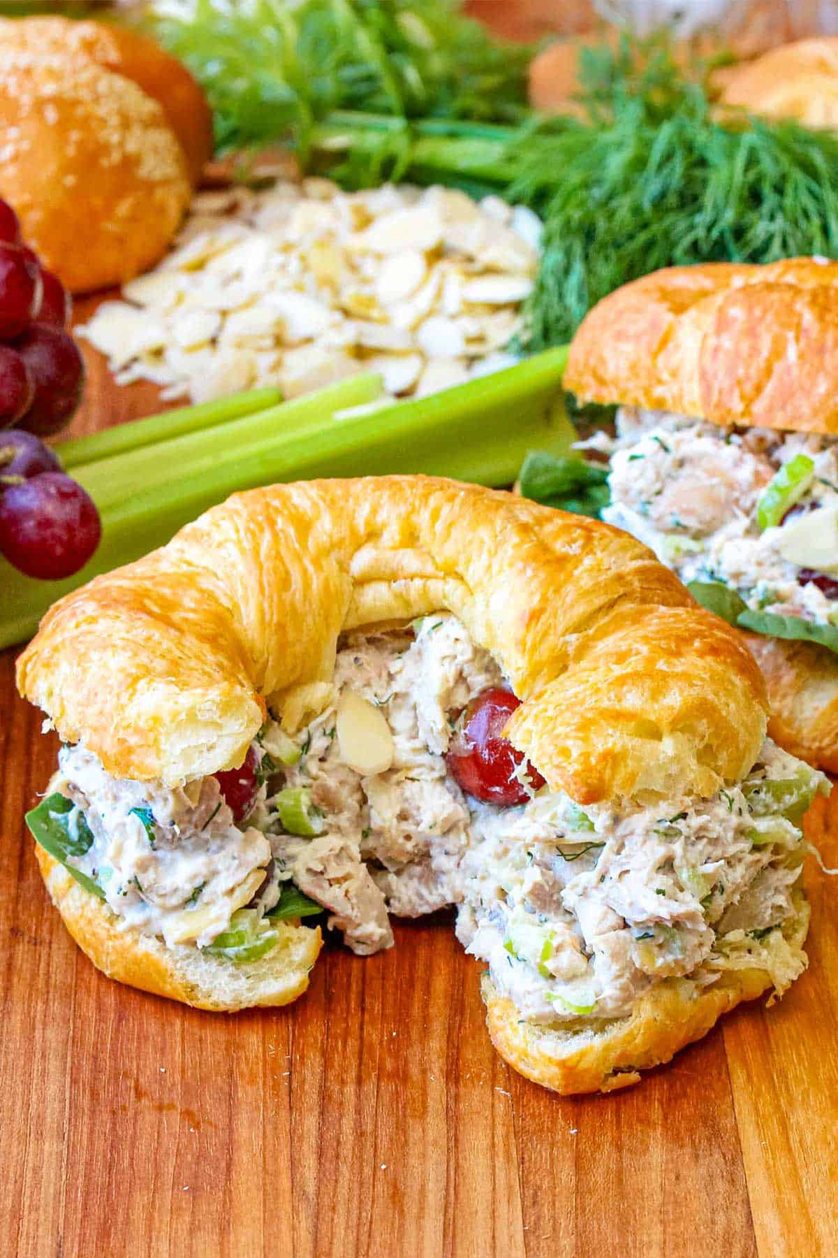 A croissant the is stuffed with Best-Ever Chicken Salad sitting on a cutting board next to another croissant also stuffed with the salad also resting near grapes, celery, and herbs.