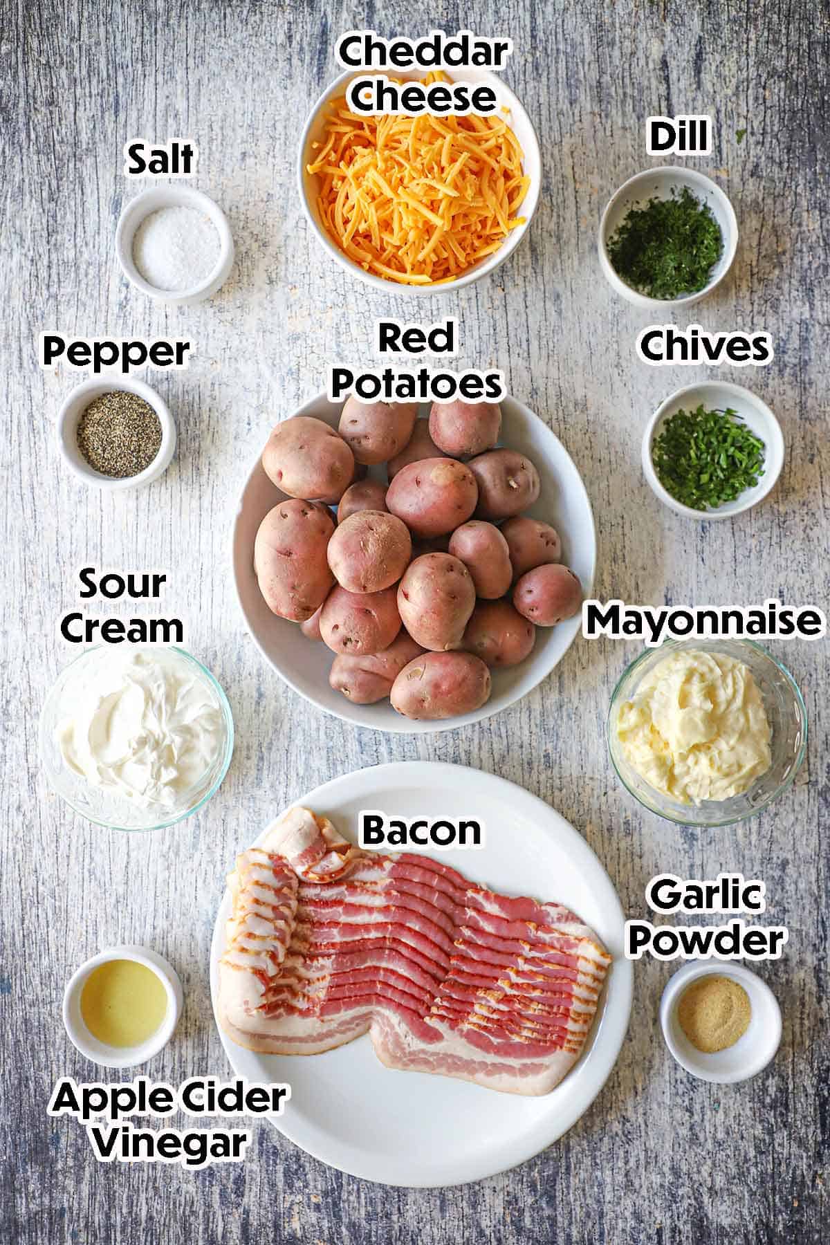 An arrangement of ingredients for creamy loaded potato salad on a grey wooden background including red potatoes, bacon, sour cream, mayonnaise, apple cider vinegar, cheese, herbs, and seasonings.