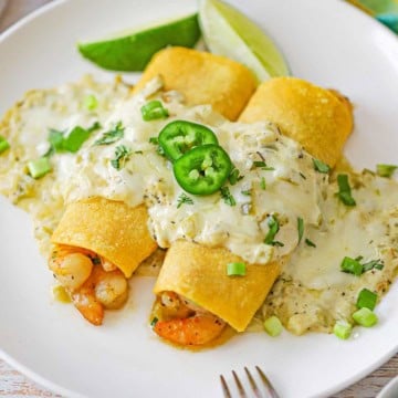 A white dinner plate filled with two creamy shrimp enchiladas that show the pink shrimp inside the rolled corn tortillas and topped with a creamy sauce and garnished with two sliced of jalapeños.