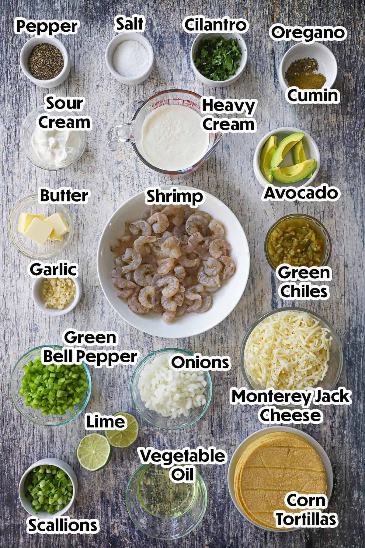 An arrangement of ingredients for Creamy Shrimp Enchiladas on a grey wooden background including raw shrimp, onions, bell pepper, scallions, lime, green chiles, cream, sour cream, butter, corn tortillas, and seasonings.