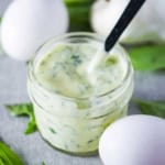 A close-up view of a small jar filled with garlic basil aioli with a small spoon inserted into it with a couple of whole eggs and fresh basil resting nearby.