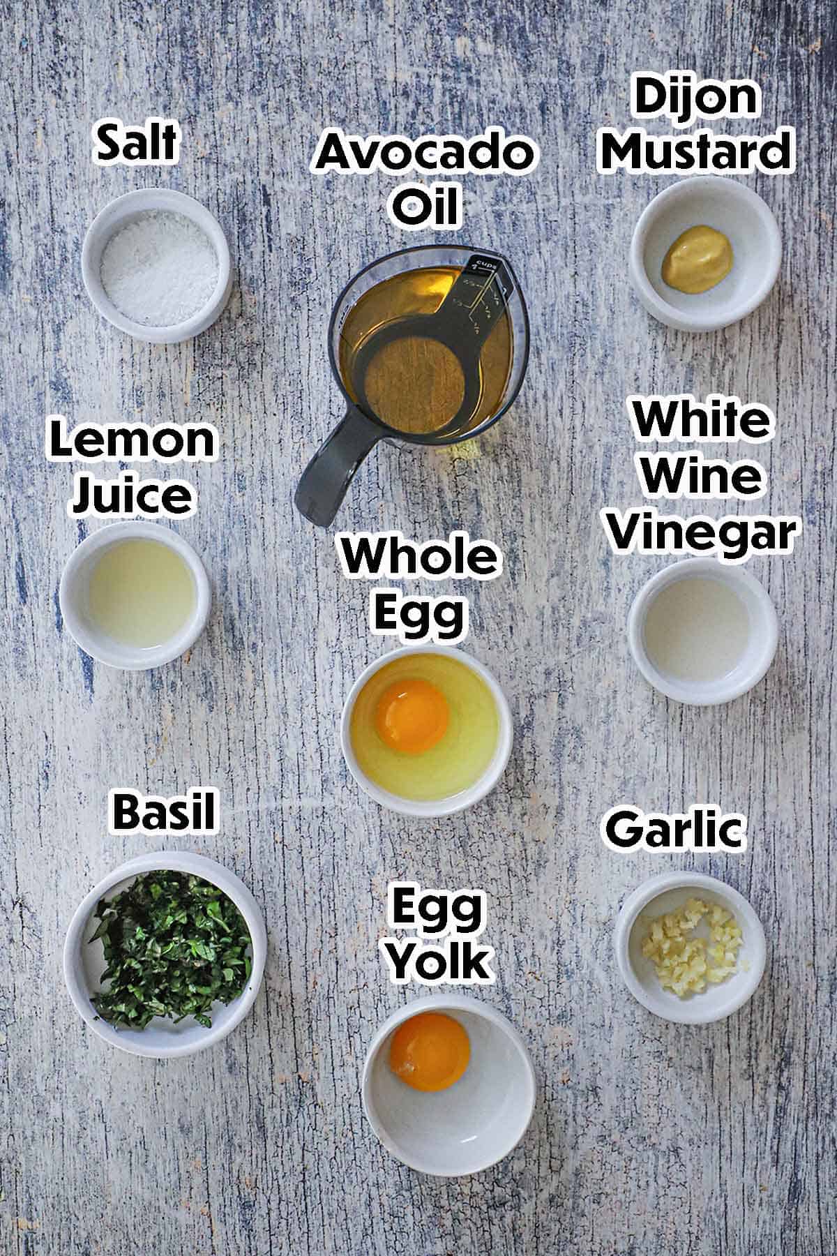 An arrangement of ingredients for garlic basil aioli on a grey wooden background including a whole egg, an egg yolk, vinegar, Dijon mustard, lemon juice, oil, garlic, basil, and salt.