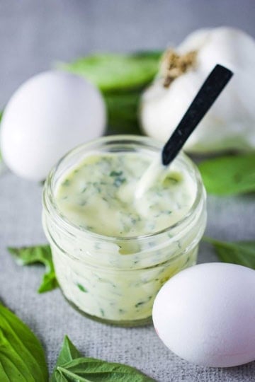 A straight-on view of a small jar filled with garlic basil aioli with a small spoon inserted into it with a couple of whole eggs and fresh basil resting nearby.